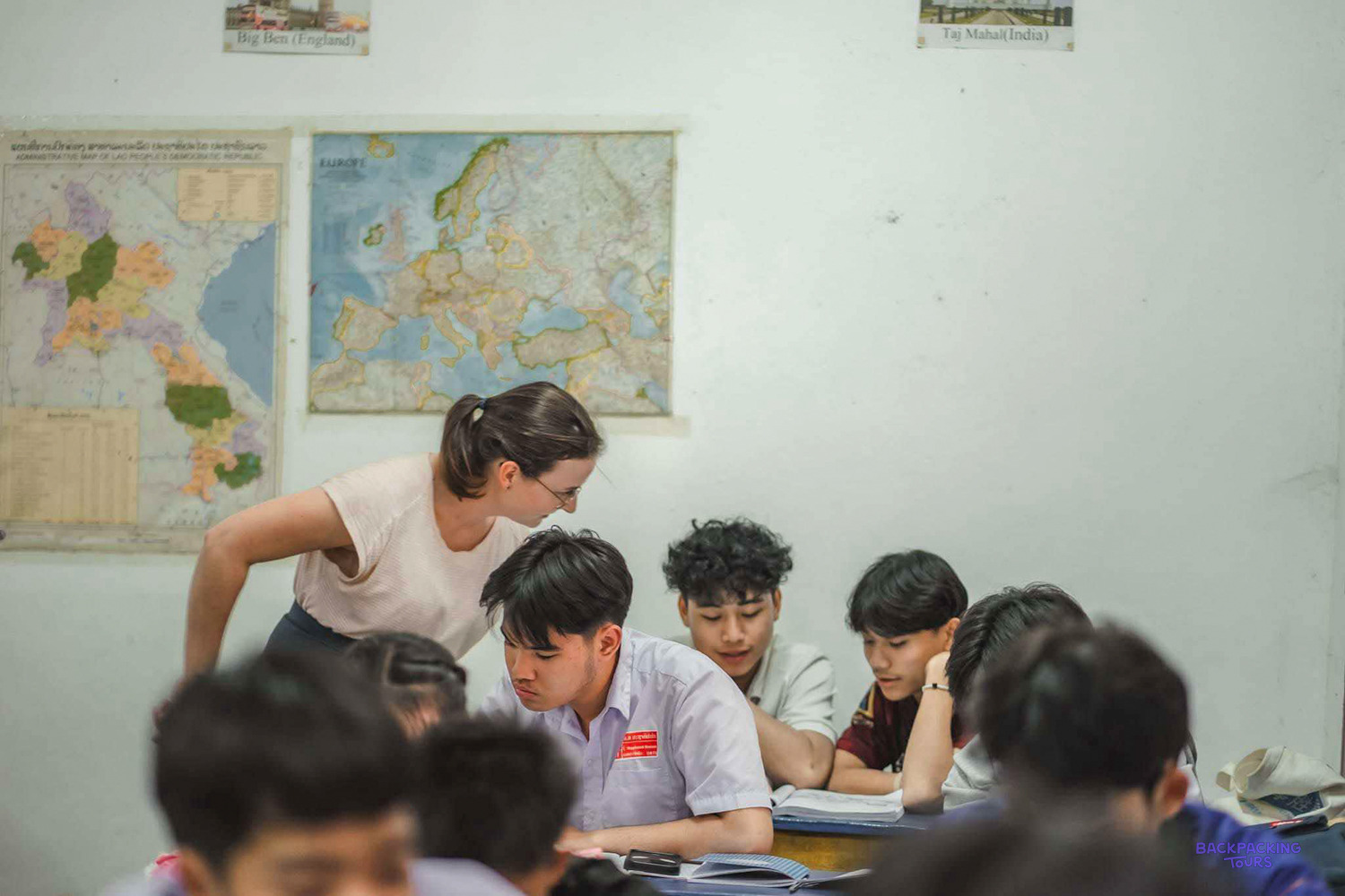 A volunteer leans in to help a group of teenage students beneath maps on the classroom wall.