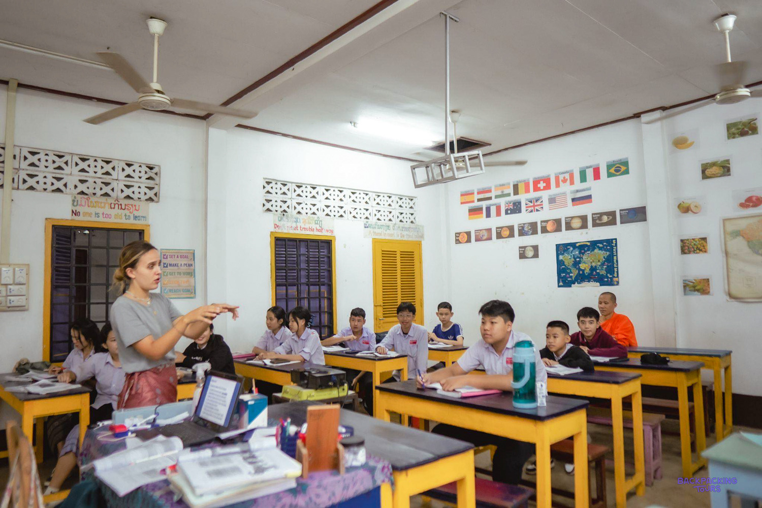 A volunteer leads an English lesson for teenage students and a young monk at yellow desks.