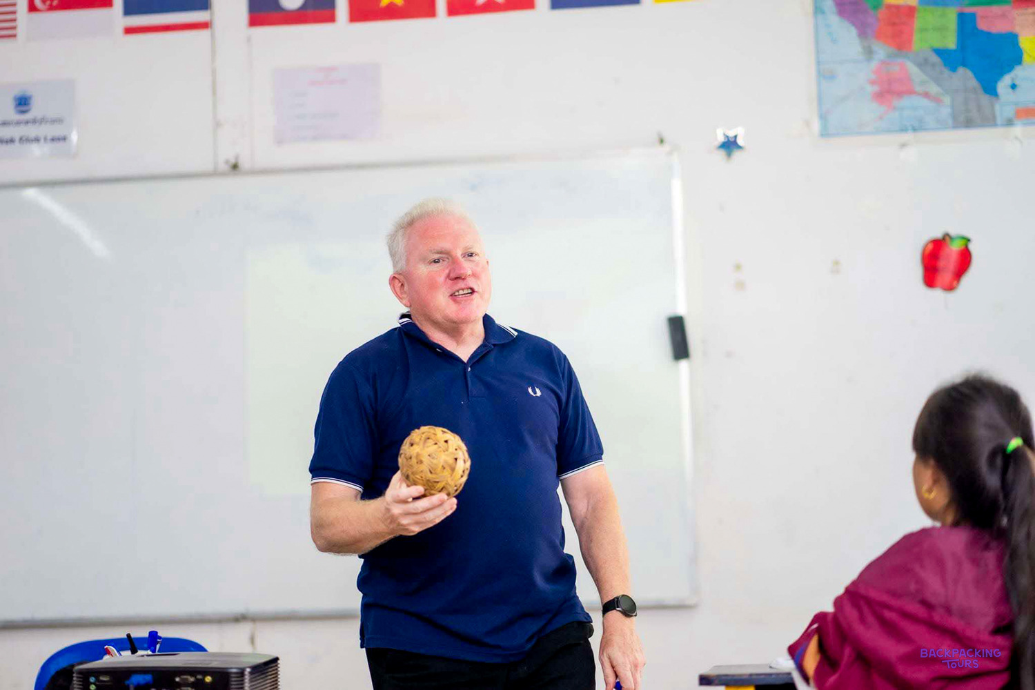 A volunteer holds up a woven rattan ball while teaching in front of a whiteboard.