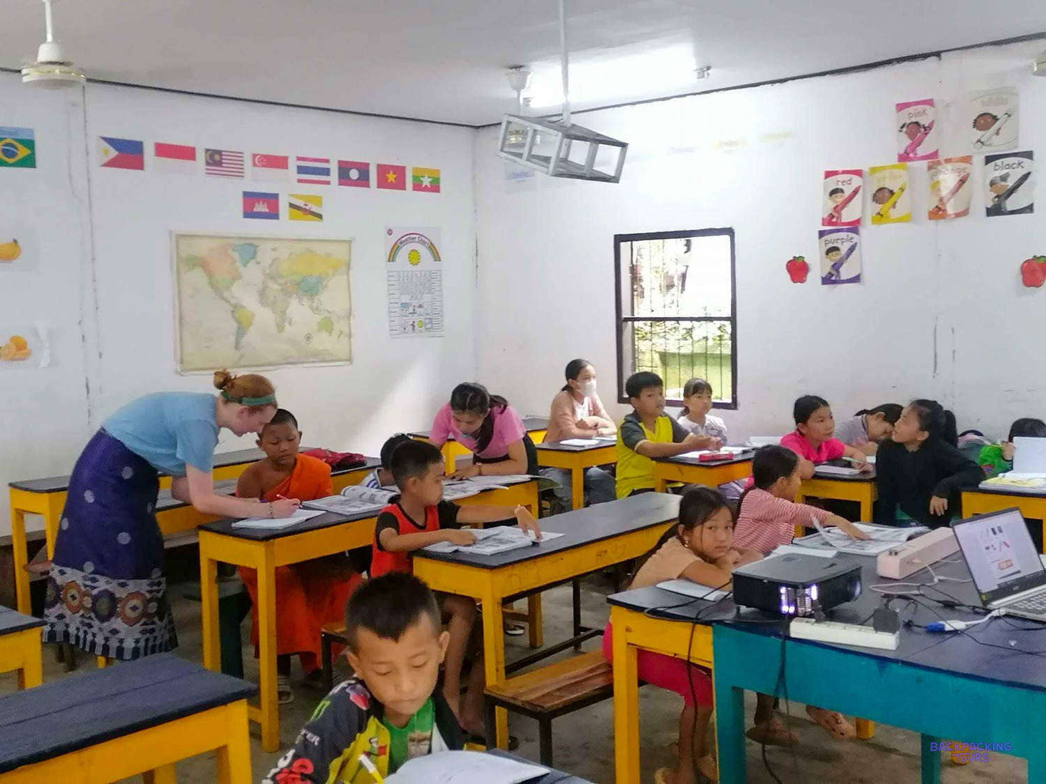 A volunteer in a Lao skirt bends to help young pupils working at yellow desks.