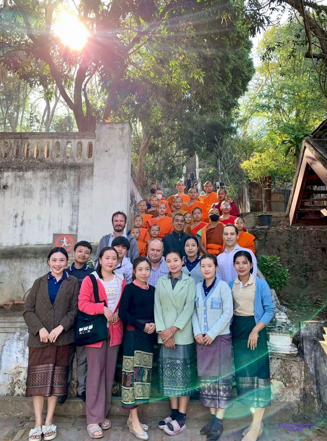 Volunteers and local people stand on a stone stairway with young monks in orange robes behind.