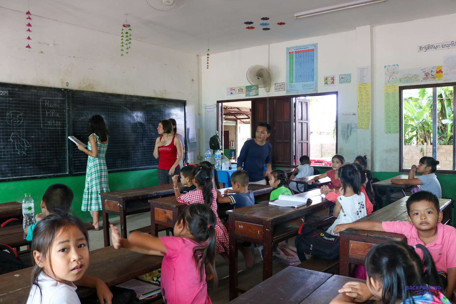 Two volunteers write on the blackboard while young pupils sit at wooden desks and look on.