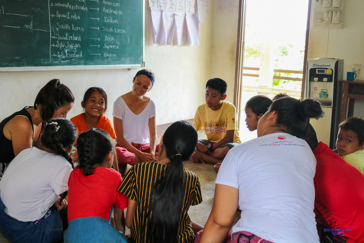 Volunteers sit on the floor in a circle with smiling Lao children during a lesson.