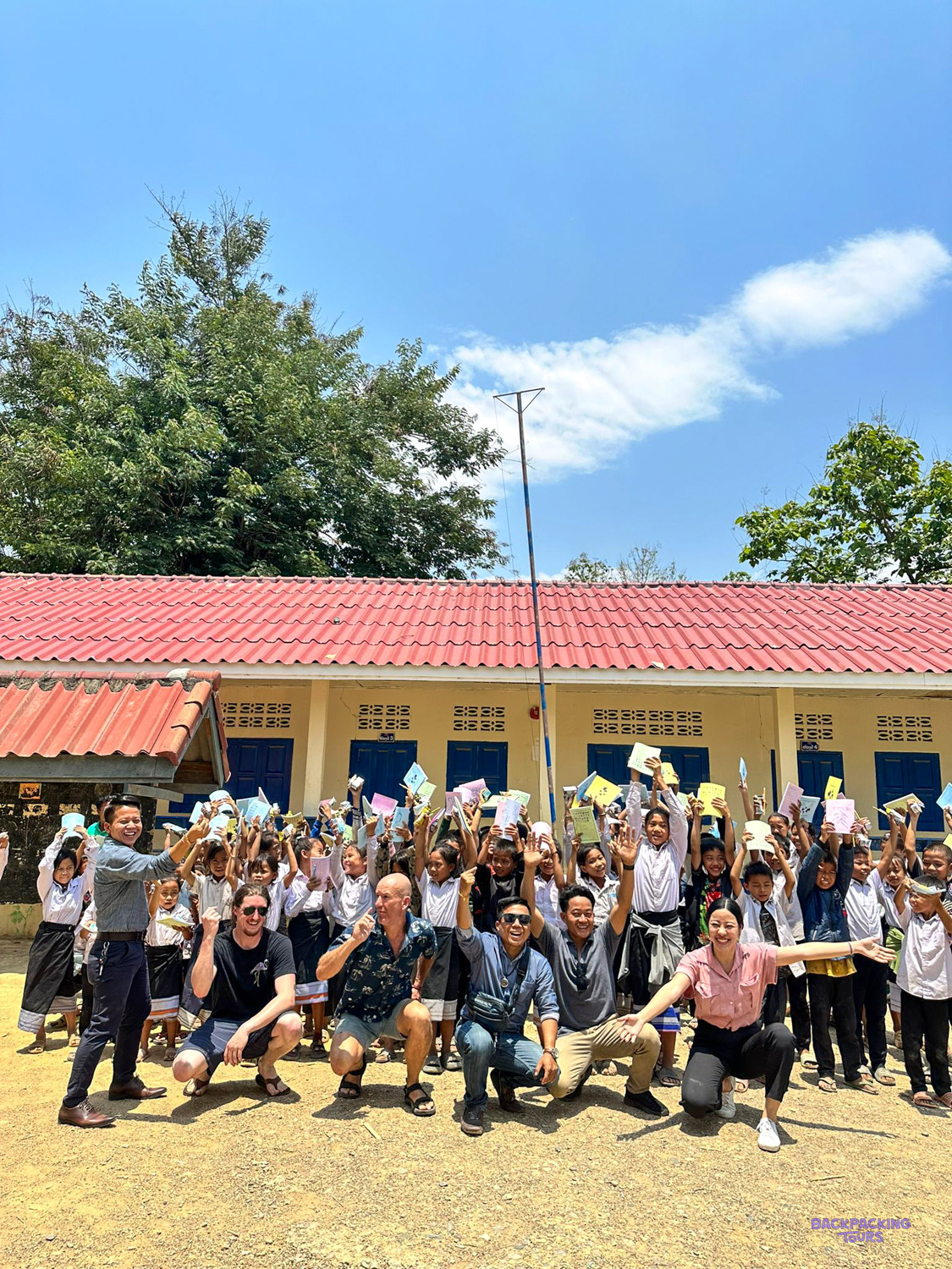 Volunteers crouch in front of cheering Lao pupils holding up new exercise books outside their school.