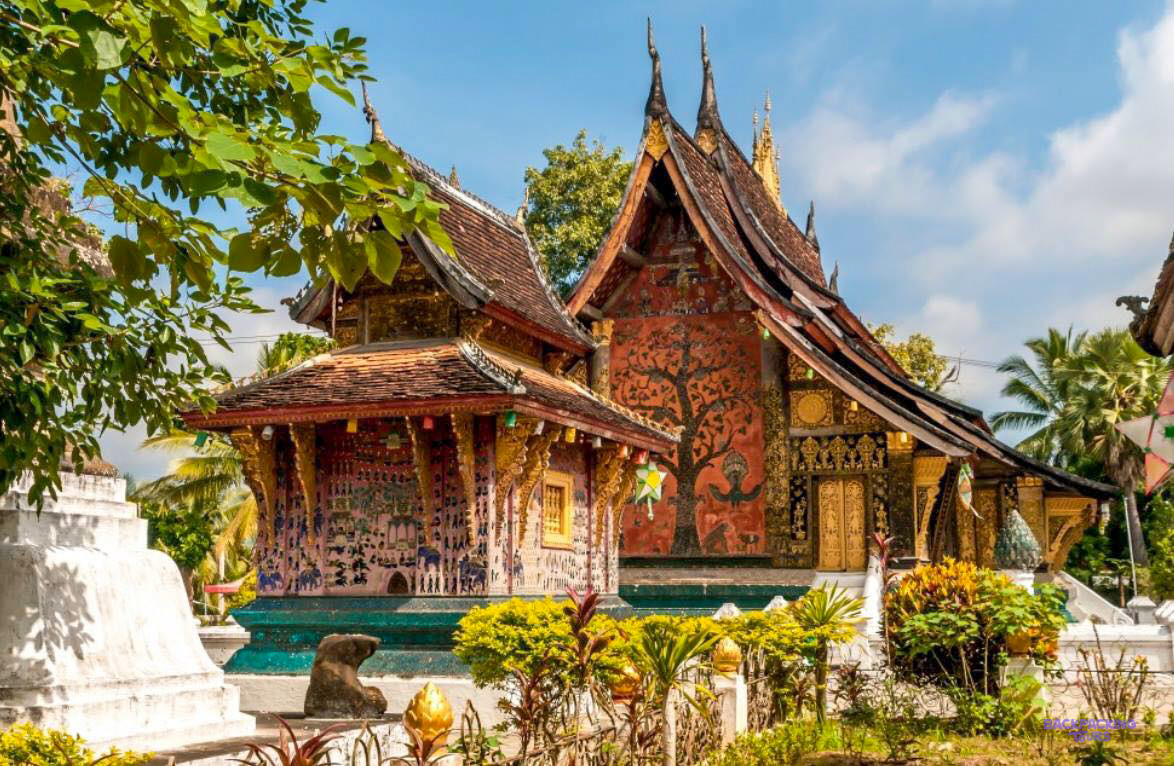 The red chapel and the tree of life mosaic at Wat Xieng Thong, framed by tropical gardens.