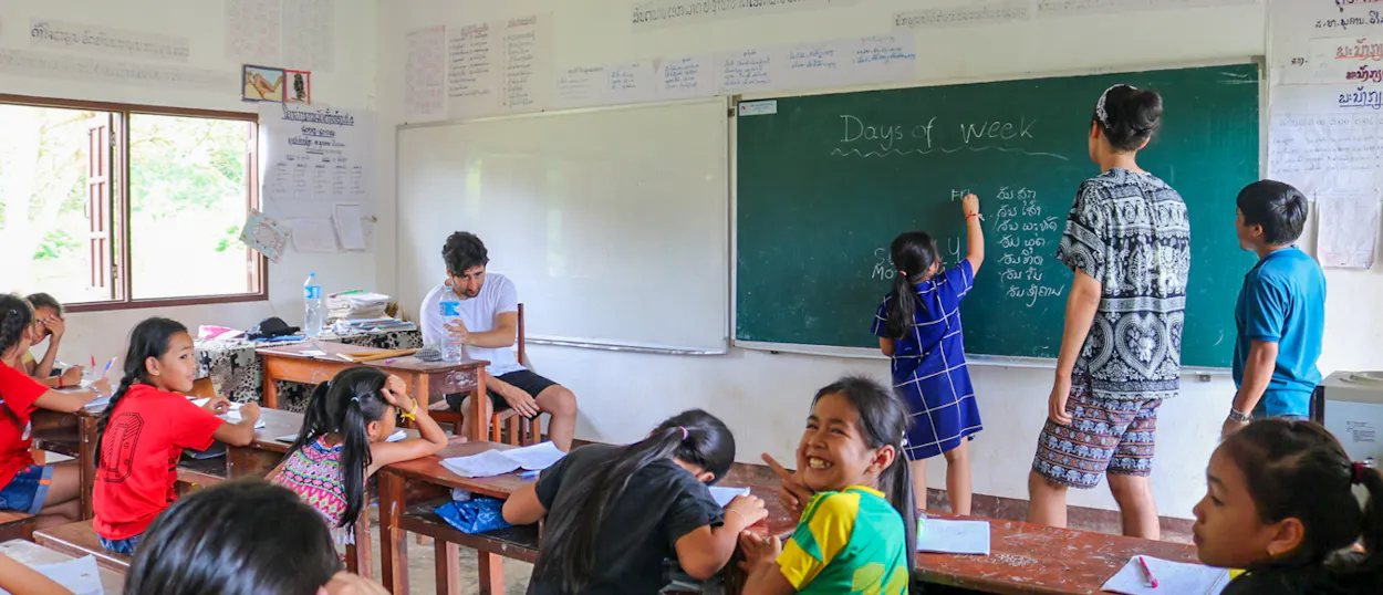 A young pupil writes on a blackboard headed Days of week, helped by a volunteer.