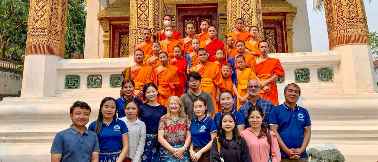 Volunteers and monks in orange robes gather on the steps of an ornate gilded temple.