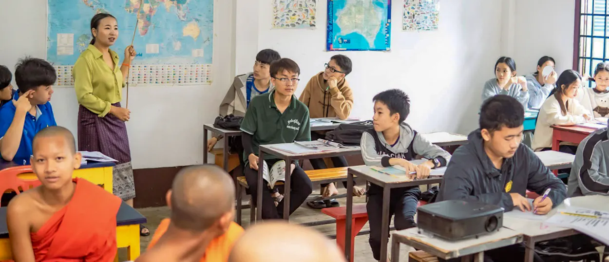 A Lao teacher stands by a world map in front of a class of students and young monks.
