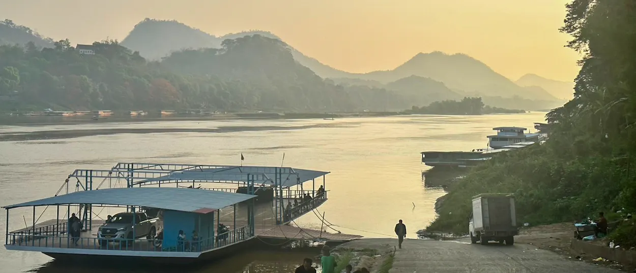 A car ferry waits at a slipway on the Mekong as mist settles over the hills beyond.