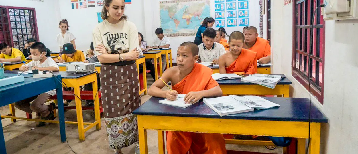 A volunteer stands between desks as novice monks in orange robes work through their English books.