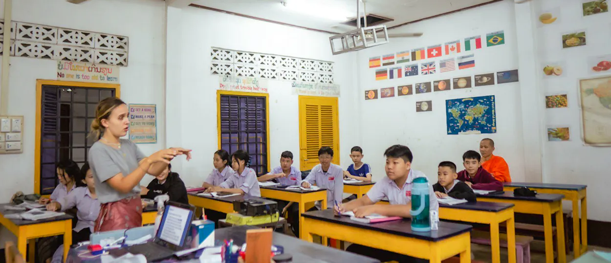 A volunteer leads an English lesson for teenage students and a young monk at yellow desks.