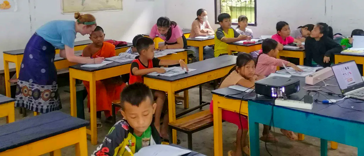 A volunteer in a Lao skirt bends to help young pupils working at yellow desks.