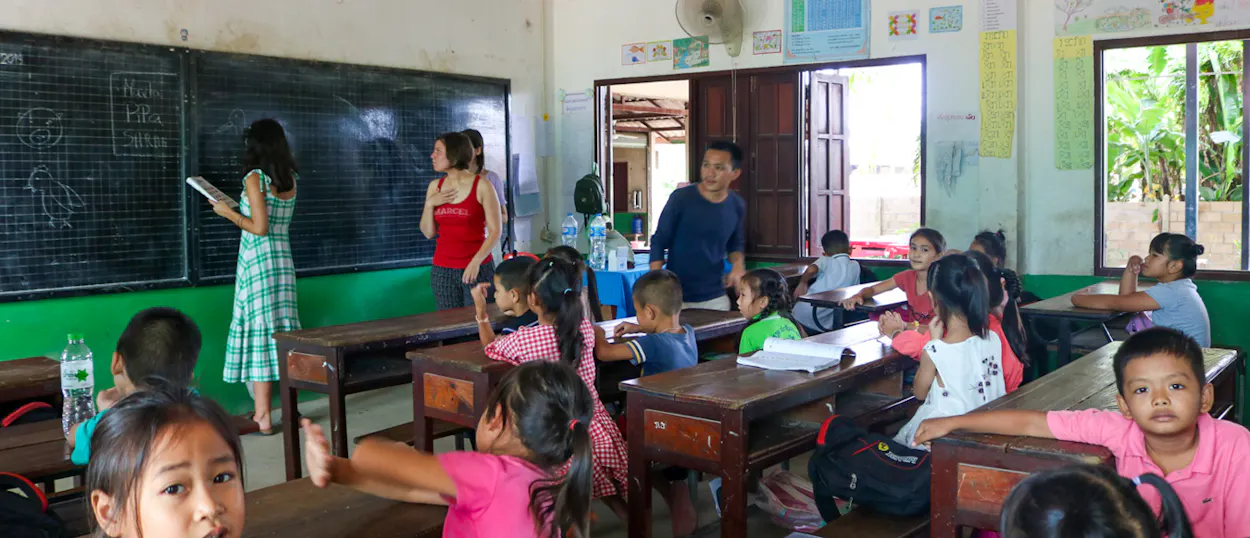 Two volunteers write on the blackboard while young pupils sit at wooden desks and look on.