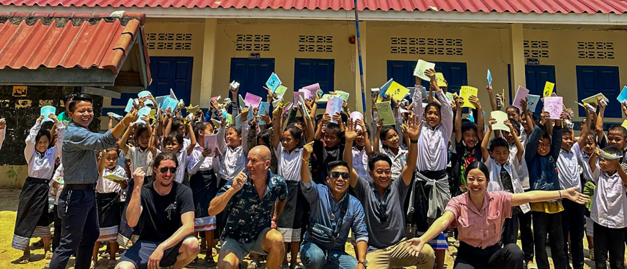 Volunteers crouch in front of cheering Lao pupils holding up new exercise books outside their school.