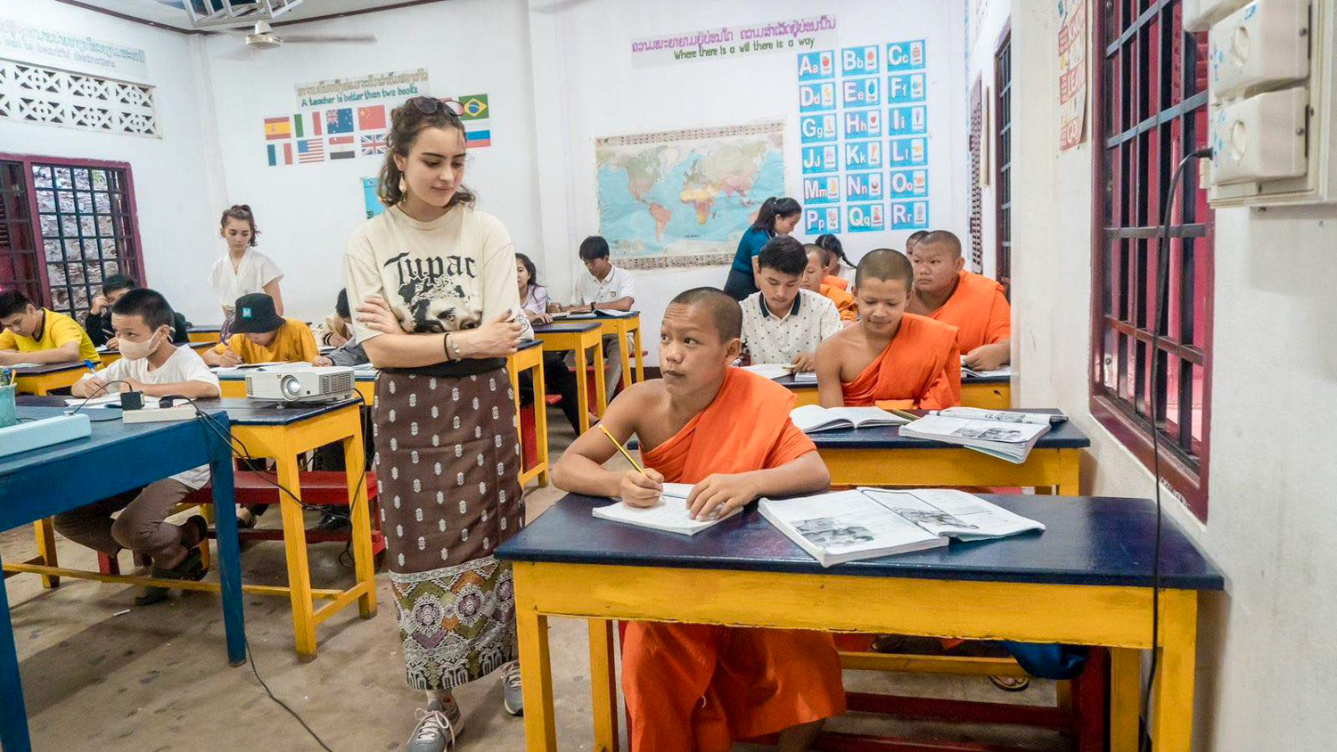 A volunteer stands between desks as novice monks in orange robes work through their English books.