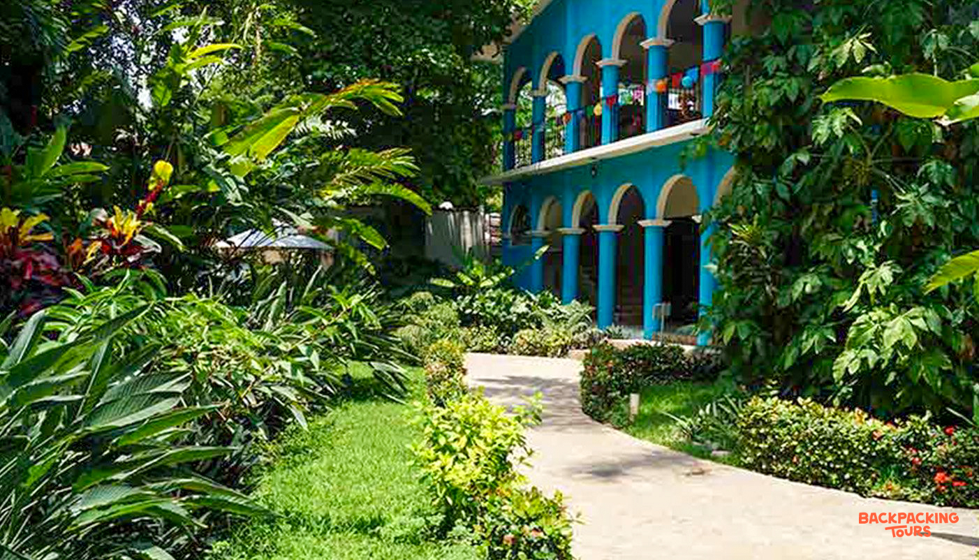 Lush tropical gardens surround a vibrant blue hotel with arched columns and balconies in Tehuantepec.