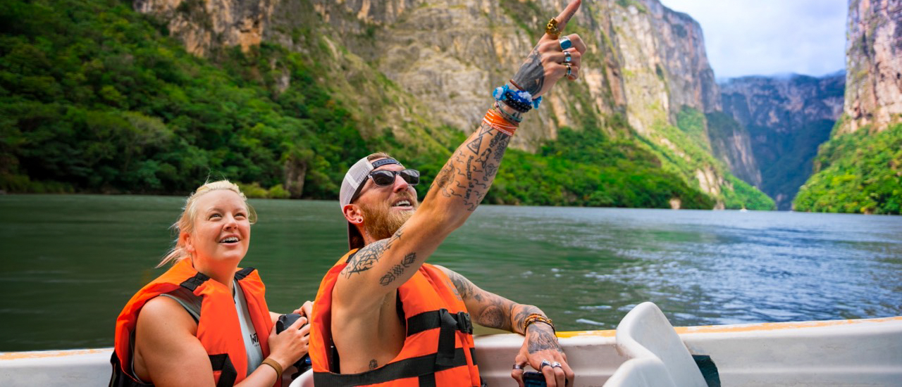 Smiling travelers in life vests on a boat cruise, pointing at towering cliffs in Sumidero Canyon.