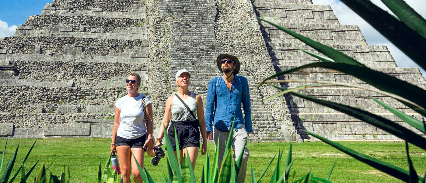 Three tourists walk on green grass towards the ancient Mayan pyramid of Chichén Itzá.