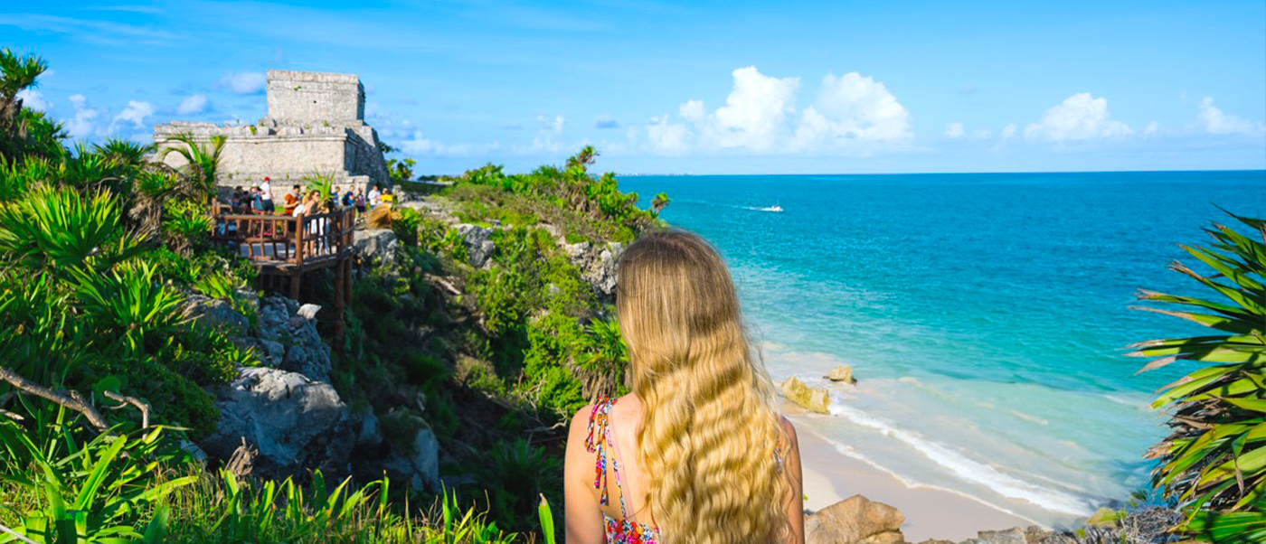 Woman admires ancient Tulum ruins and the stunning turquoise Caribbean Sea from a lush cliff.