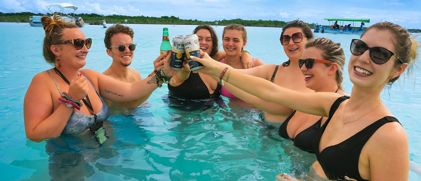 Seven friends smiling and toasting with drinks in the clear blue waters of Bacalar Lagoon.