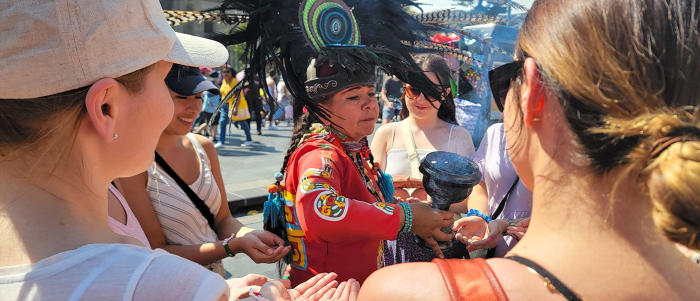 A person in a feathered headdress performs a traditional ritual for a group of tourists in Mexico City.
