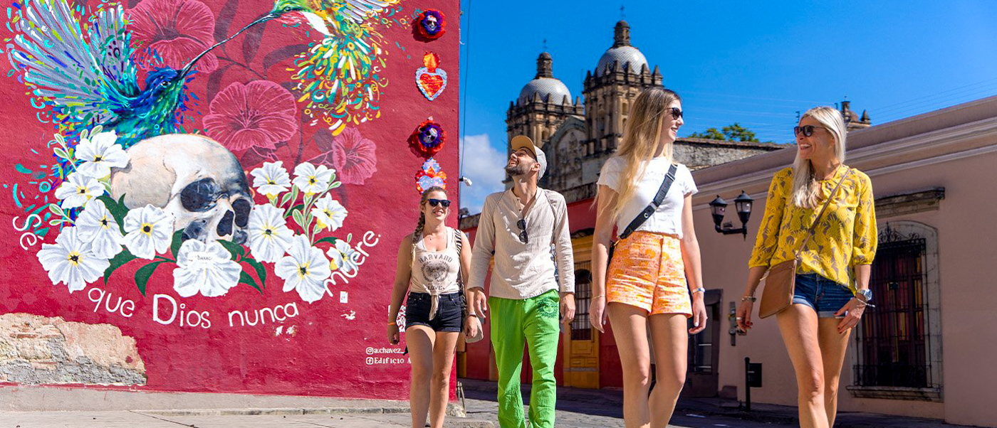 Four backpackers walk past a vibrant mural and historic cathedral in sunny Oaxaca, Mexico.