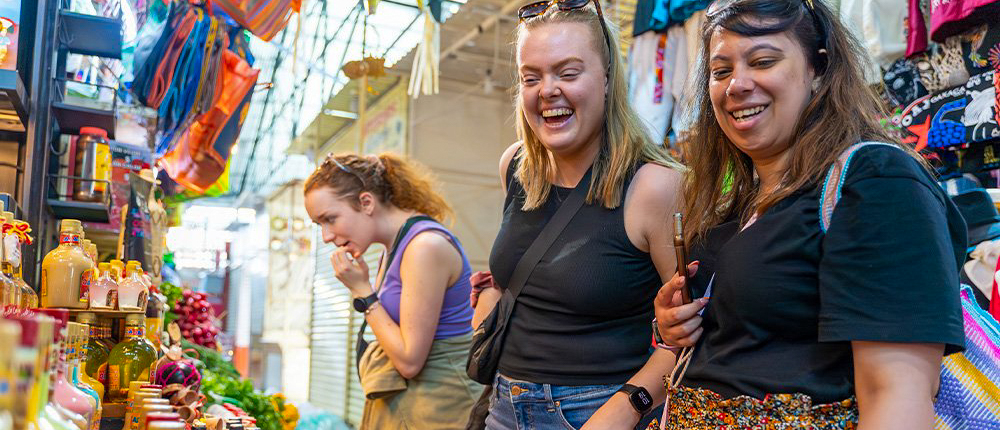 Happy travelers laughing and examining colorful bottles at a lively market stall in Tehuantepec.