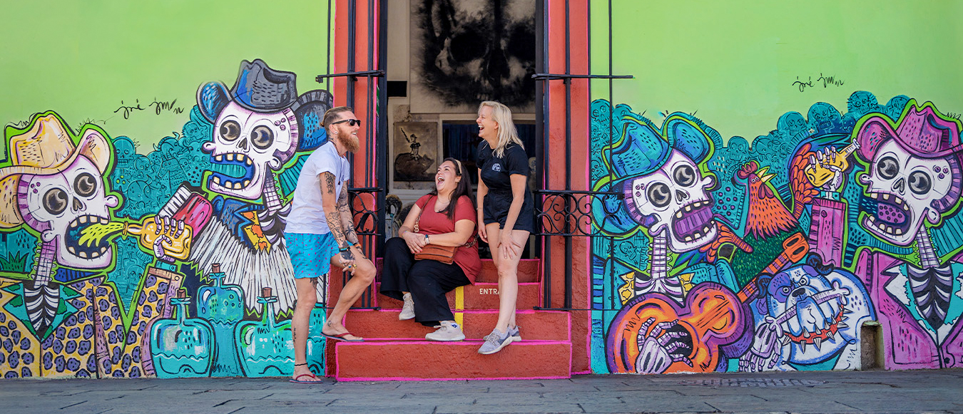 Travelers laughing on red steps in front of a vibrant Day of the Dead mural in Mexico City.