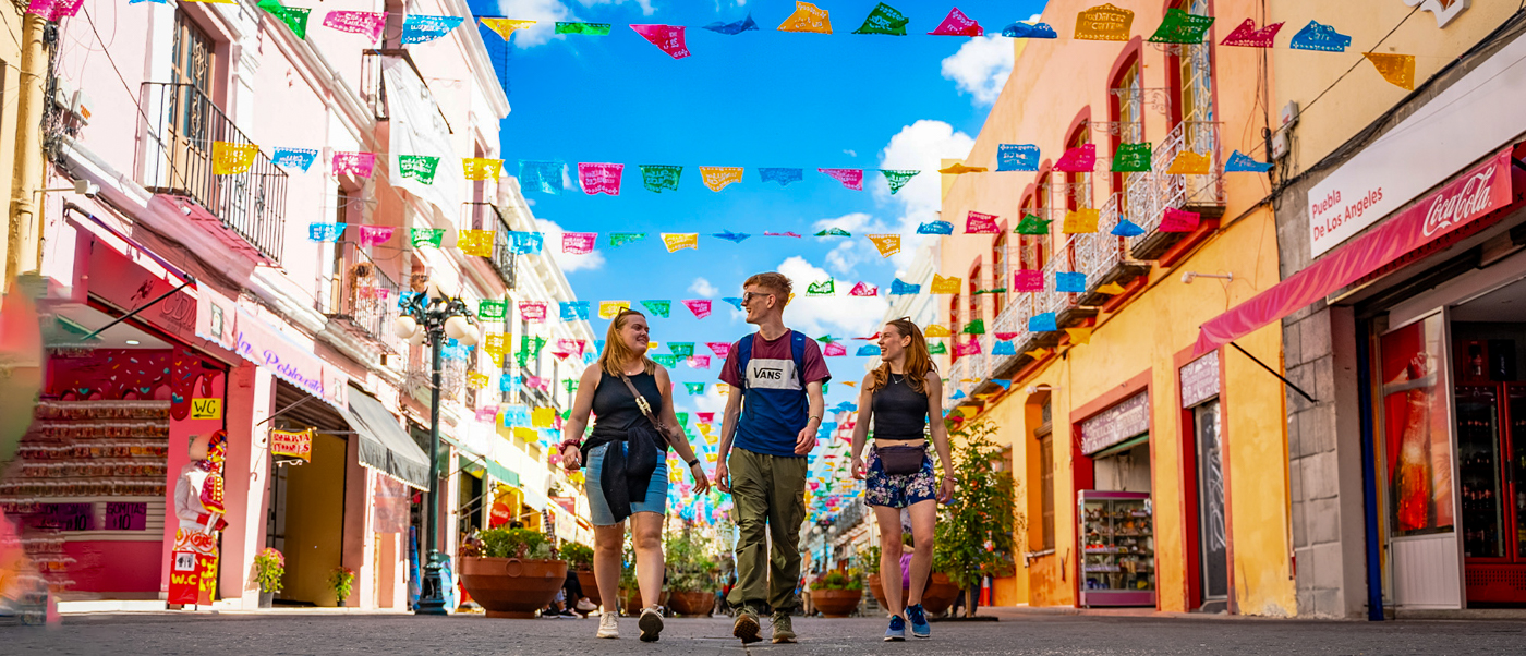 Three friends stroll a lively street in Oaxaca, adorned with colorful papel picado under a bright blue sky.