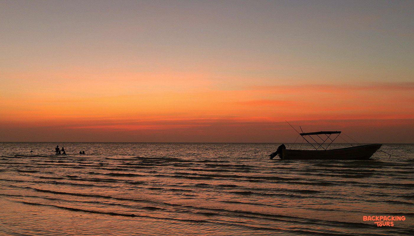 A vibrant sunset paints the sky over Playa del Carmen beach, with swimmers and a boat.