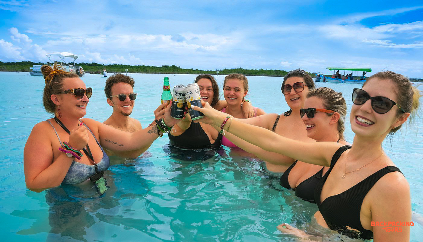 Happy backpackers toast with beers in the clear turquoise water of Bacalar Lagoon, Mexico.