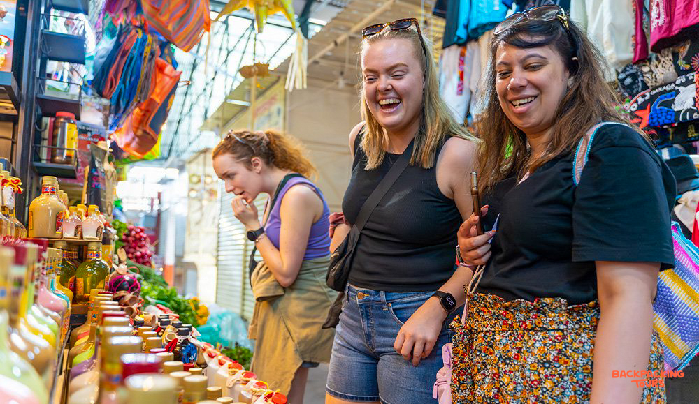 Three happy women browse colorful bottles and textiles at a vibrant market in Tehuantepec, Mexico.