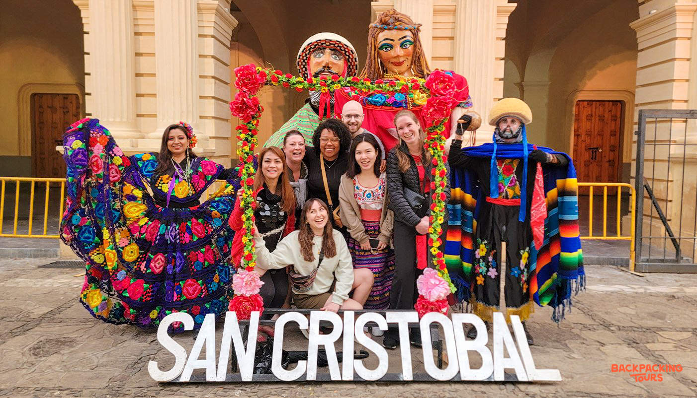 A joyful group of backpackers pose with vibrant traditional figures and a large San Cristobal sign.