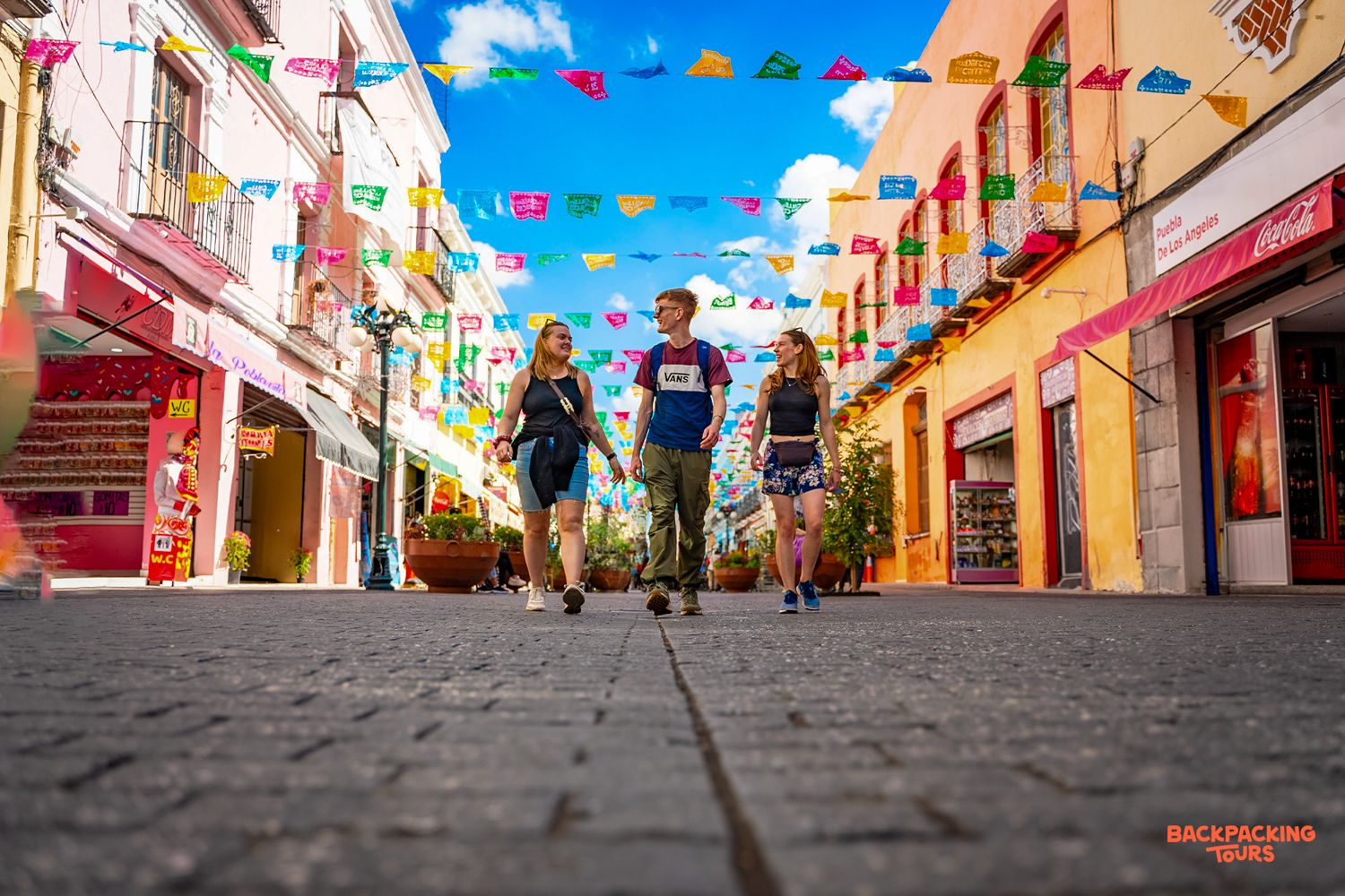Three friends stroll a vibrant Oaxaca street, enjoying the colorful buildings and festive papel picado banners.