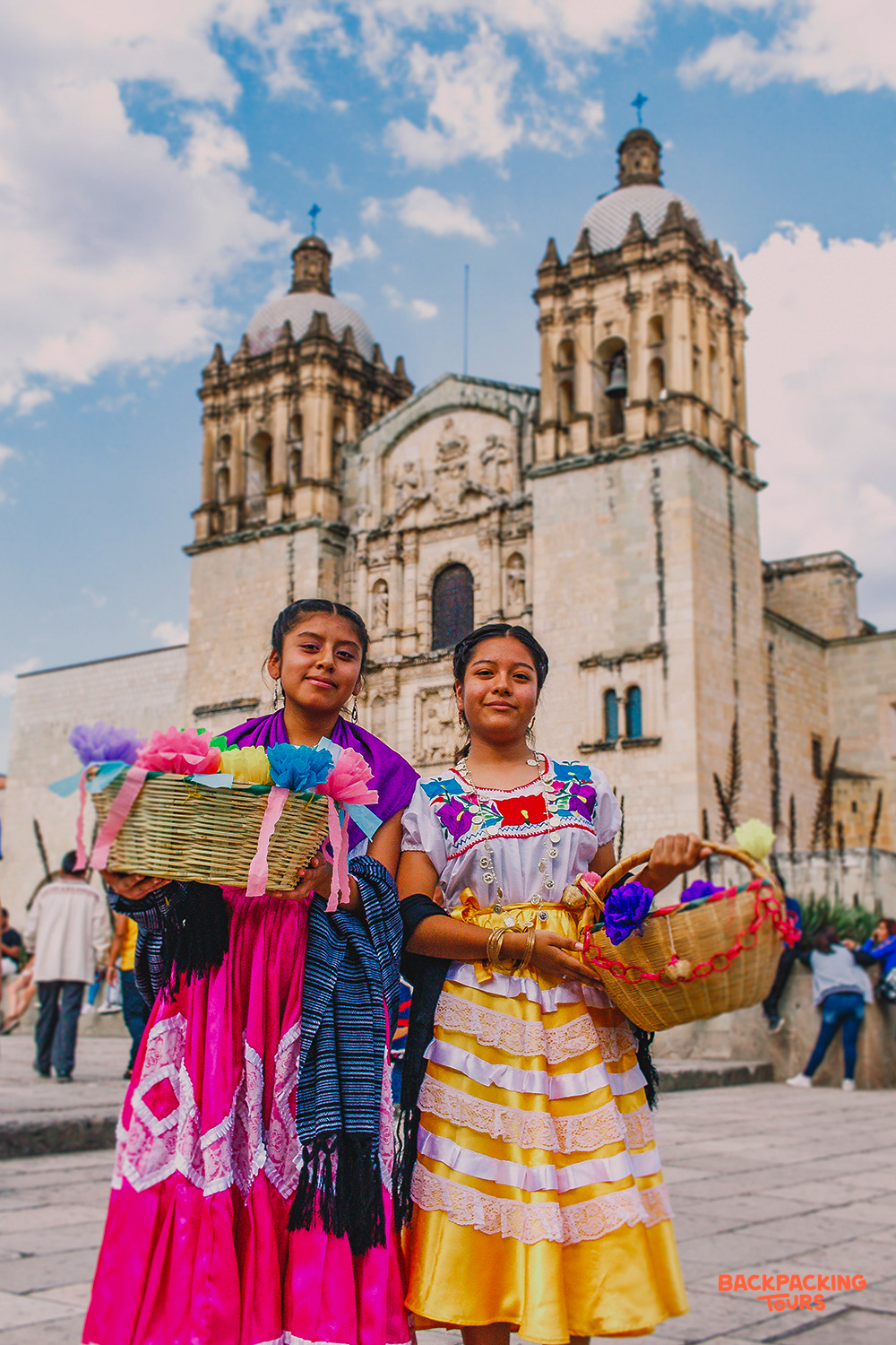 Two girls in traditional dresses hold baskets of colorful crafts in front of a grand cathedral in Oaxaca.