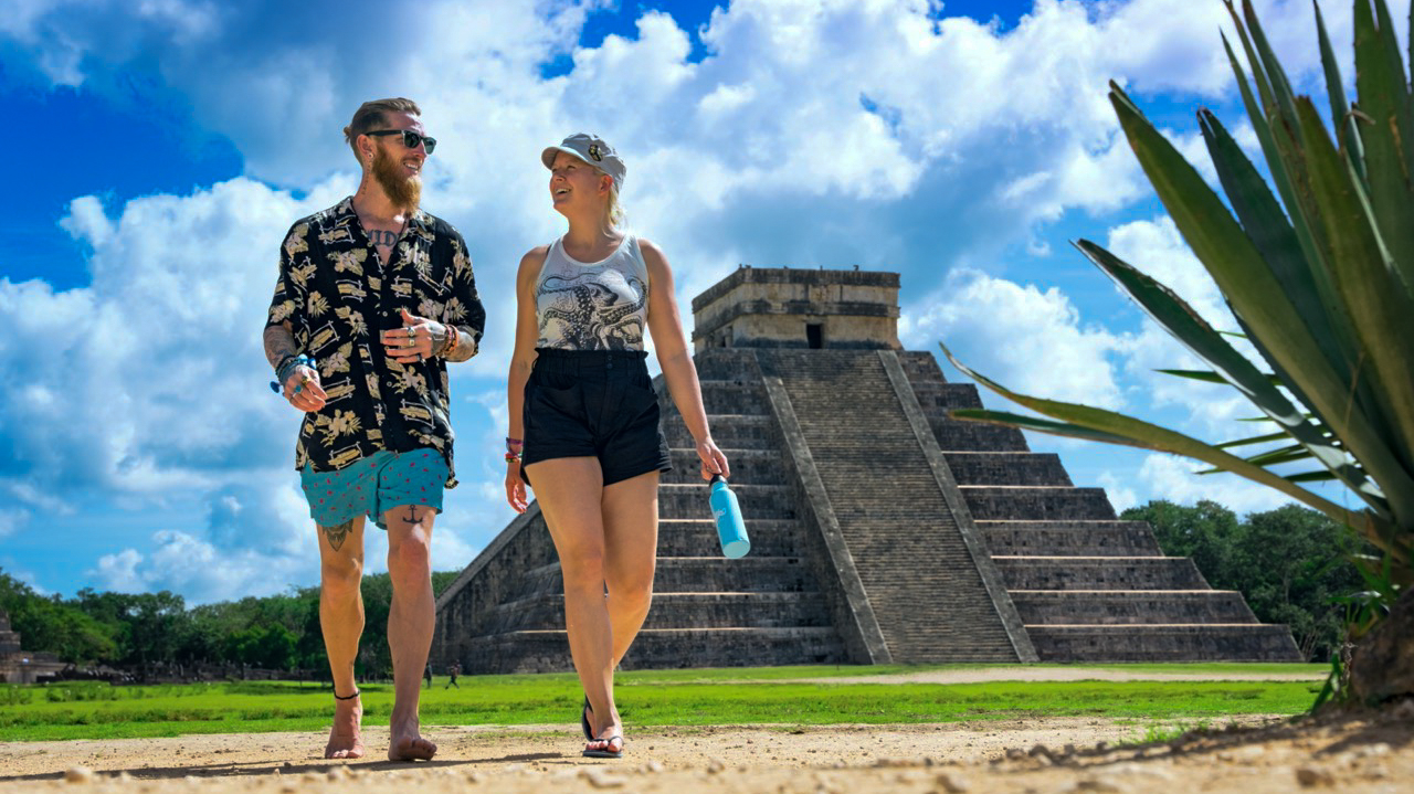 A couple walks past the ancient Chichen Itza pyramid under a bright blue sky.