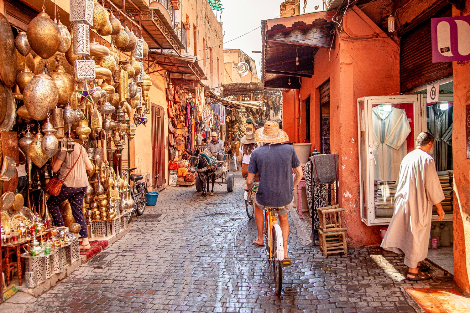 A person cycles through a vibrant, narrow market street in Marrakech, lined with brassware and shops.
