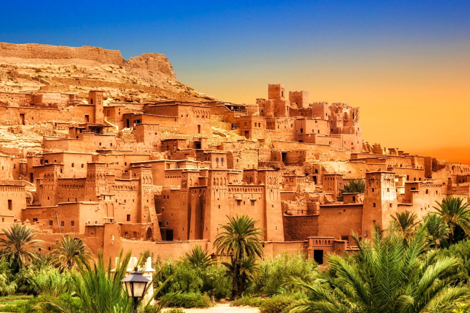 Ancient fortified village of Ait Benhaddou in Morocco, with palm trees under a colorful sky.