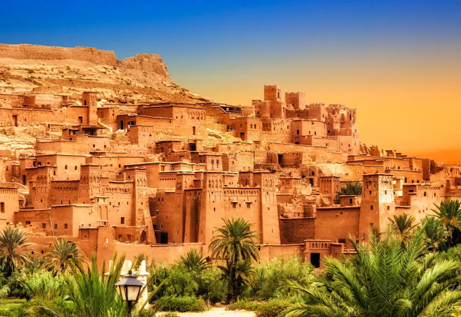 Ancient fortified village of Ait Benhaddou in Morocco, with palm trees under a colorful sky.