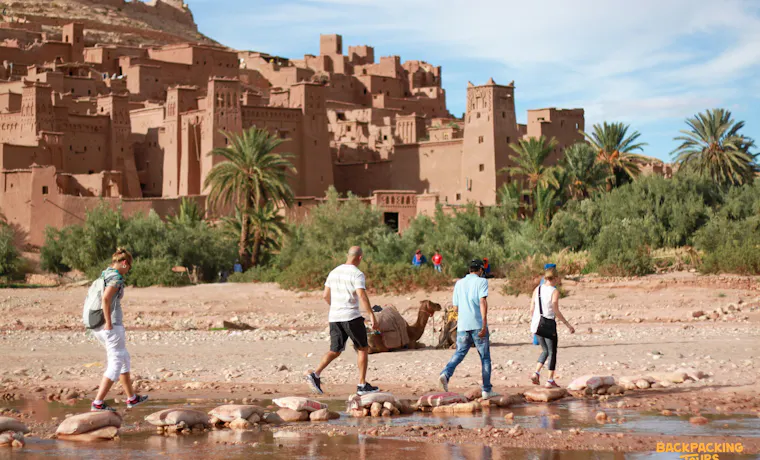 Four travelers cross a river on stepping stones with the historic Ait Ben Haddou Kasbah in Morocco.