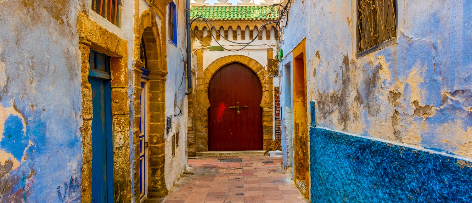 A vibrant, narrow alley in Essaouira's historic medina, featuring blue walls and a large arched red door.
