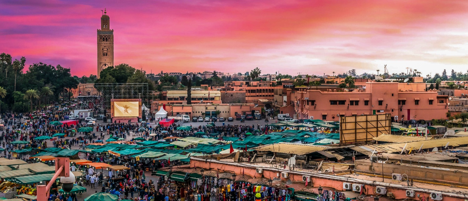 A vibrant market square in Marrakech at sunset, with a tall minaret and many bustling stalls.
