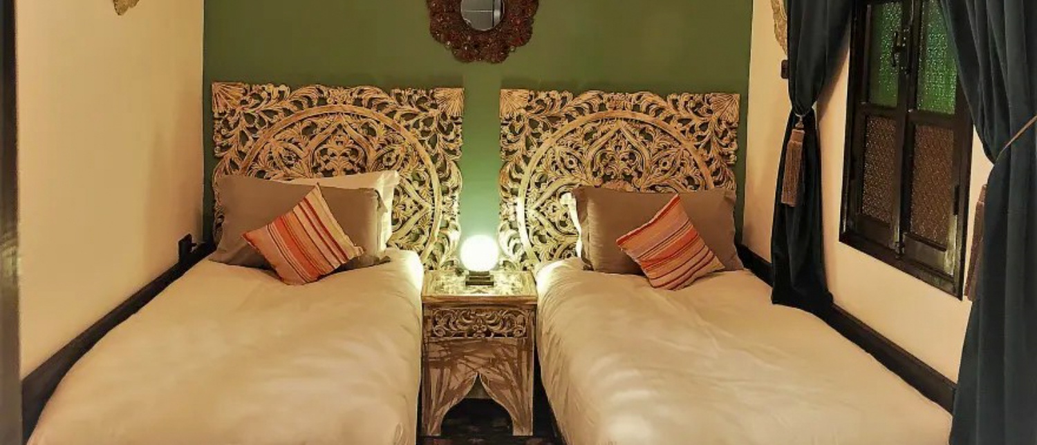 A welcoming twin bedroom with ornate wooden headboards and a warm lamp in a Marrakech riad.
