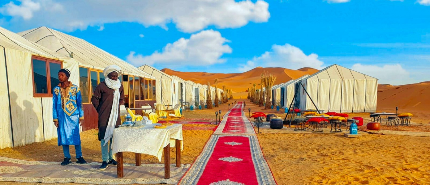 Two men in traditional attire at a desert camp with white tents and red carpet in Merzouga.