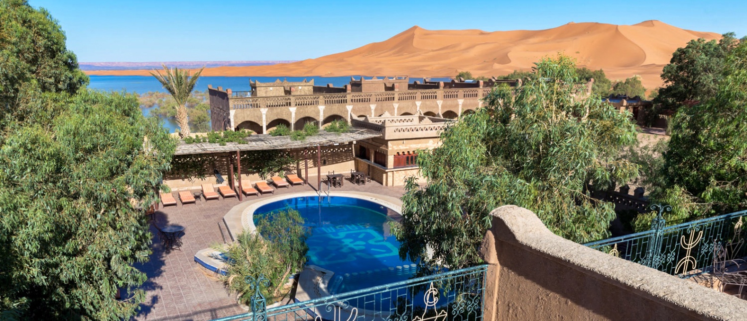 Moroccan kasbah hotel with a blue pool and desert lake, backed by Merzouga's vast sand dunes.