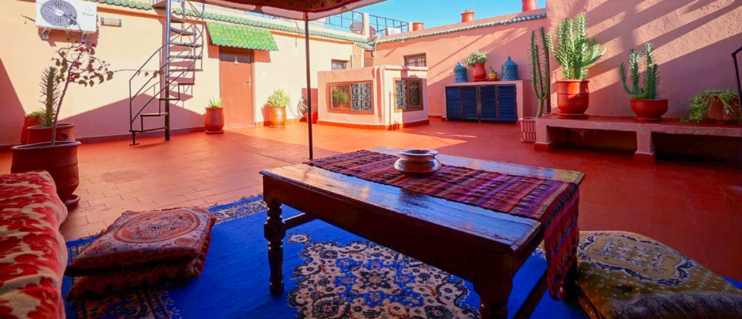 A vibrant rooftop terrace in Ouarzazate with colorful rugs, cushions, and potted cacti under a blue sky.