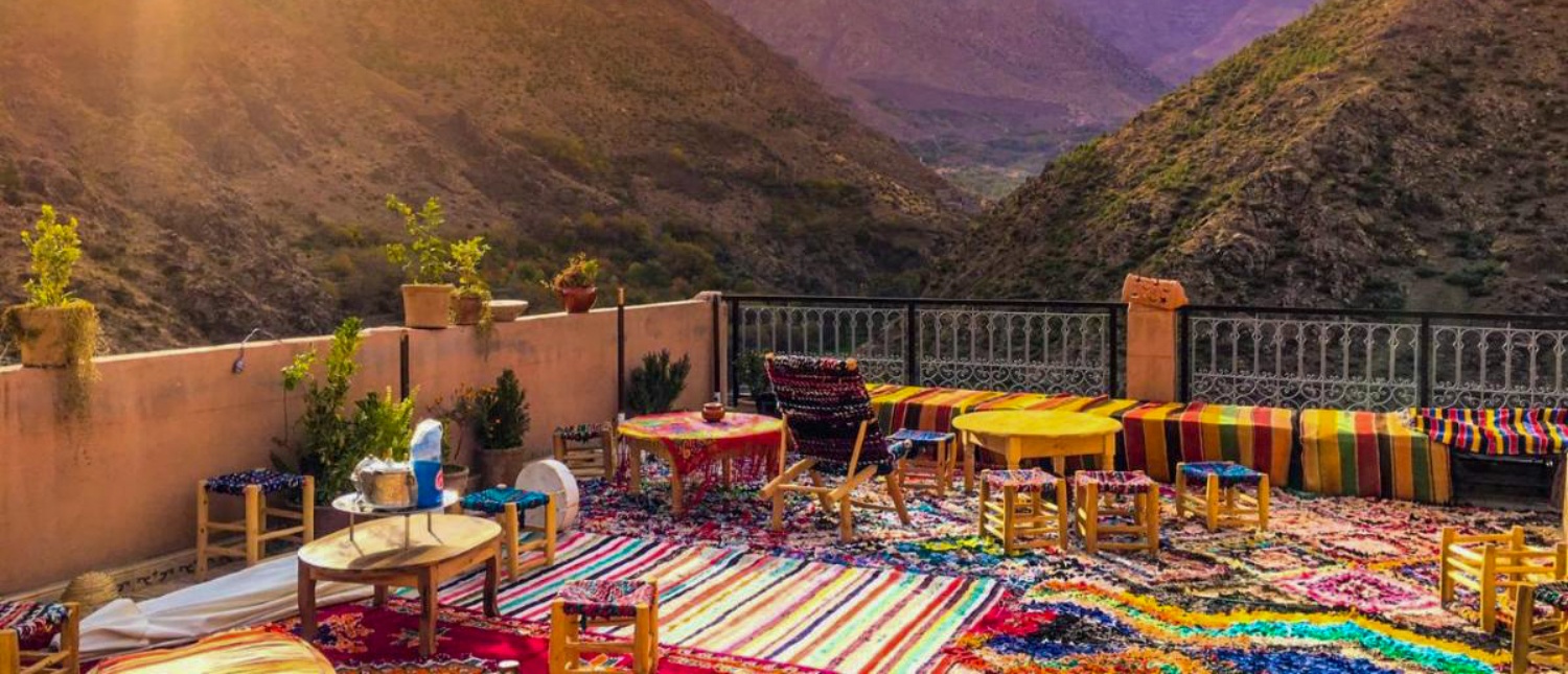 A vibrant rooftop terrace in Imlil, Morocco, with colorful rugs, low tables, and stunning mountain views.