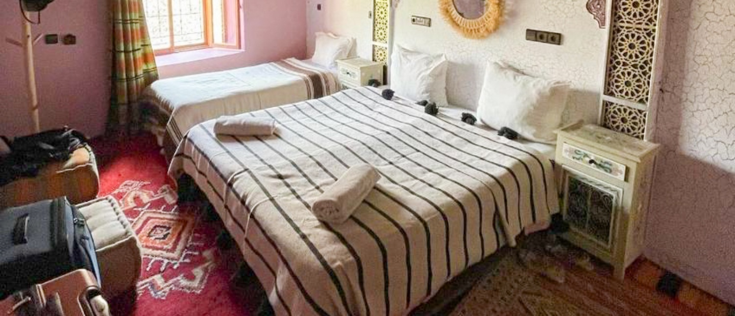 Inviting Moroccan bedroom with two beds, striped blankets, a red patterned rug, and luggage.