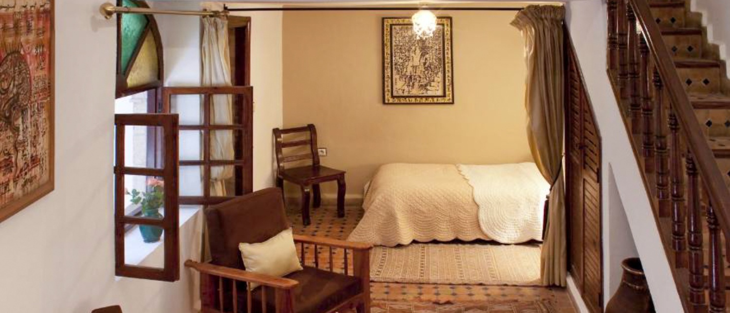 A warm guesthouse room in Essaouira, Morocco, with a bed, wooden furniture, and carved staircase.