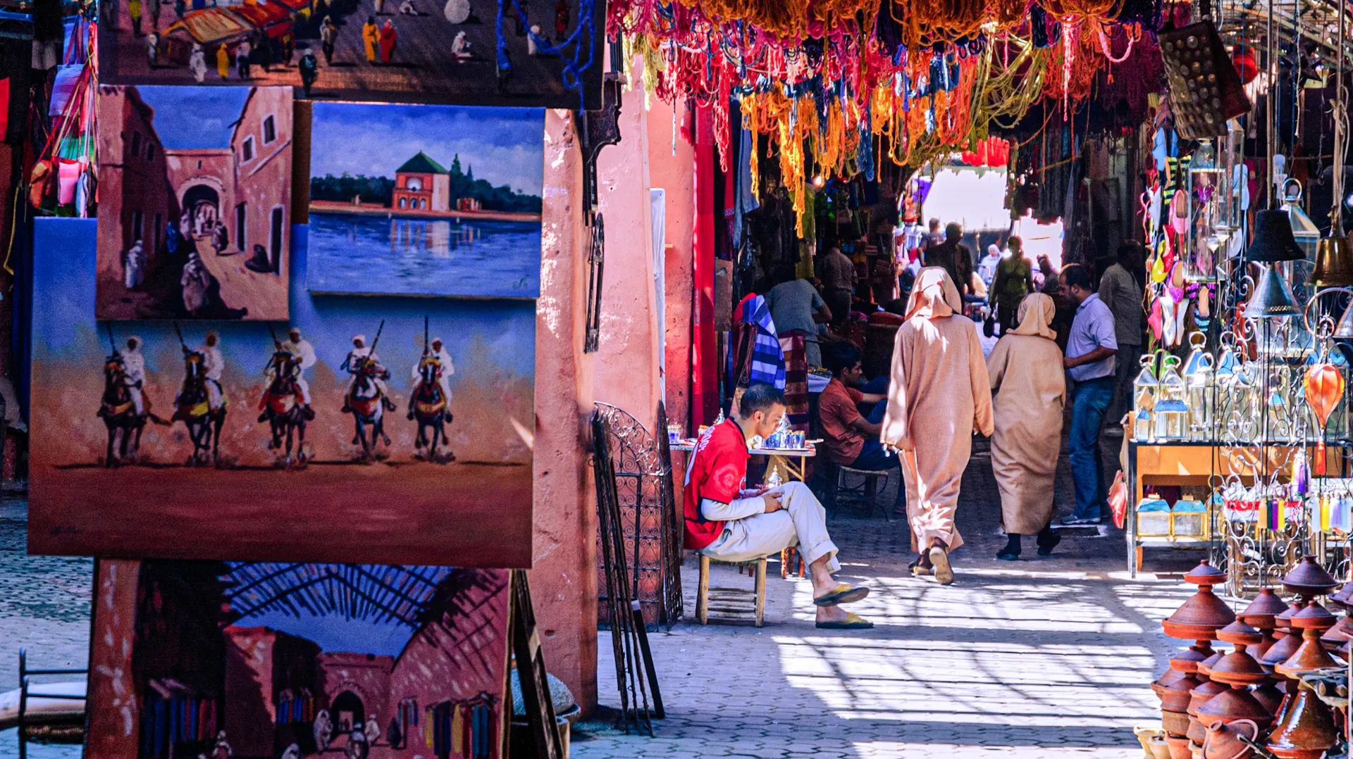 A bustling Moroccan market street with colorful textiles overhead, paintings on display, and people walking.